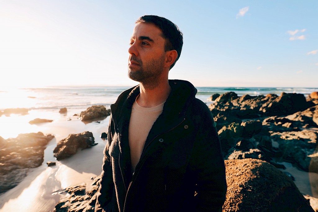adam gokmen at the beach with rocks and the ocean behind him looking towards the sun